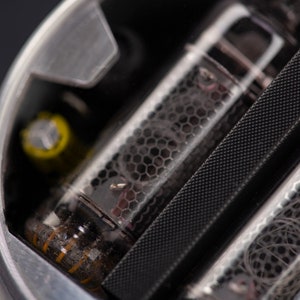 May include: Close-up of a silver-colored electronic component with glass tubes. The tubes have a honeycomb-like pattern and internal wiring. A yellow cylindrical component is visible. The background is dark.