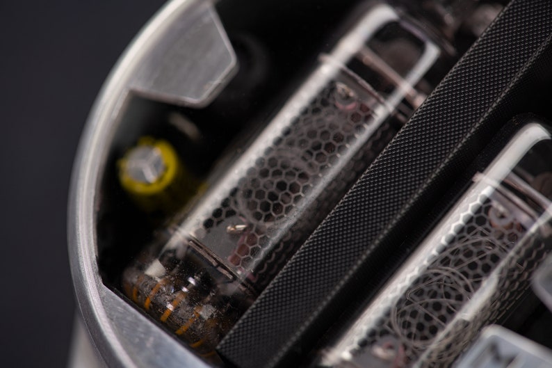 May include: Close-up of a silver-colored electronic component with glass tubes. The tubes have a honeycomb-like pattern and internal wiring. A yellow cylindrical component is visible. The background is dark.