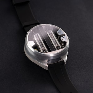 May include: A futuristic wristwatch with a silver case and a black rubber strap. The watch face is transparent, revealing two vertical tubes. The watch has two buttons labeled "H" and "M". The watch is on a black surface.