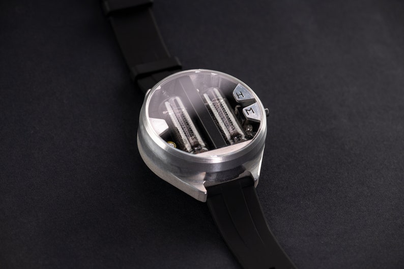 May include: A futuristic wristwatch with a silver case and a black rubber strap. The watch face is transparent, revealing two vertical tubes. The watch has two buttons labeled "H" and "M". The watch is on a black surface.