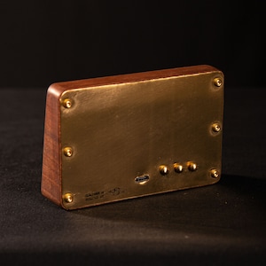 May include: A rectangular wooden device with a brass faceplate, secured by brass screws. The device has a USB port and three small buttons. The text "DESIGNED BY MEC GEAR" is visible. The device is on a black surface.
