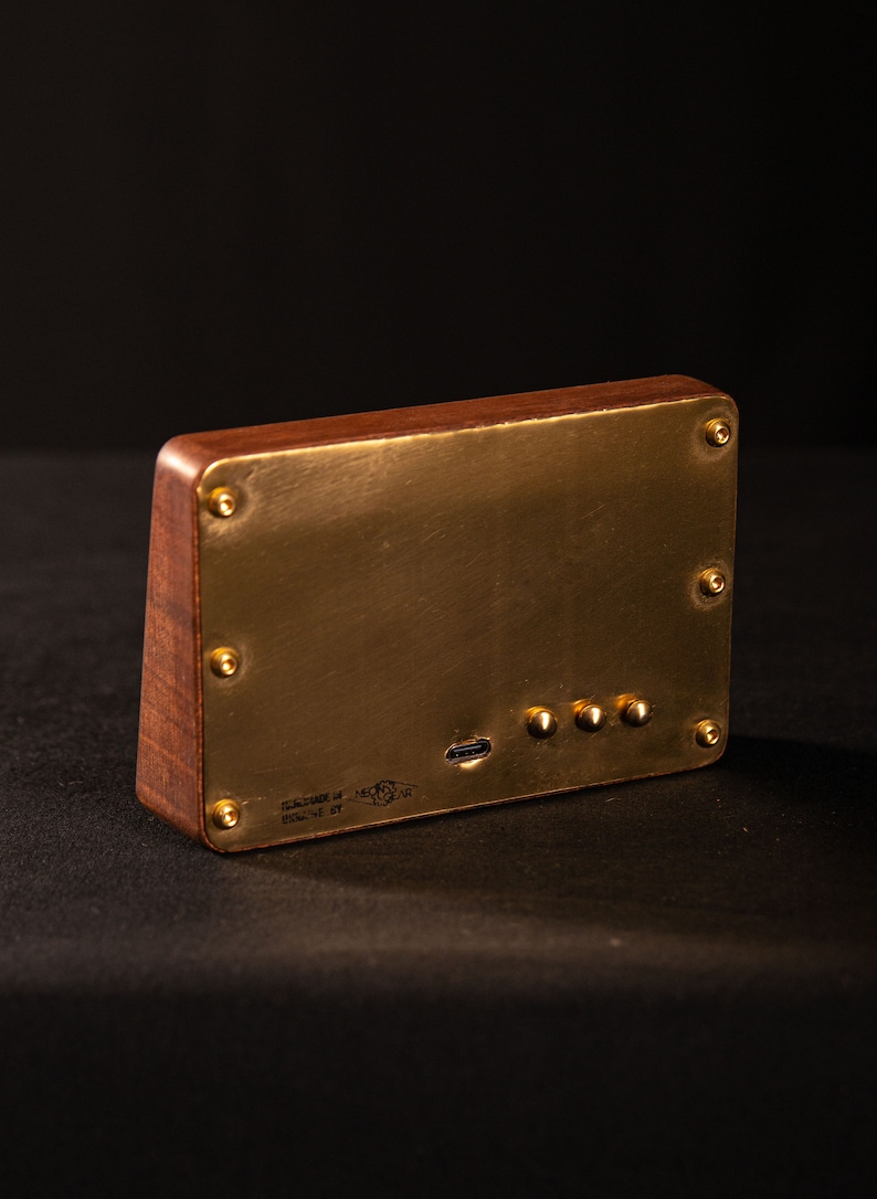 May include: A rectangular wooden device with a brass faceplate, secured by brass screws. The device has a USB port and three small buttons. The text "DESIGNED BY MEC GEAR" is visible. The device is on a black surface.