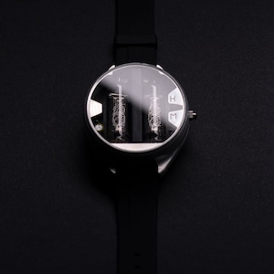 May include: A black wristwatch with a round, silver-colored face and a black rubber strap. The watch face has a clear cover, revealing two vertical tubes with intricate internal structures. The letters "H" and "M" are visible on the face.