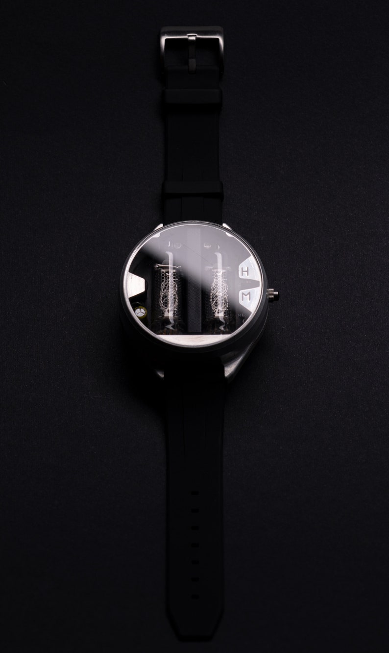 May include: A black wristwatch with a round, silver-colored face and a black rubber strap. The watch face has a clear cover, revealing two vertical tubes with intricate internal structures. The letters "H" and "M" are visible on the face.