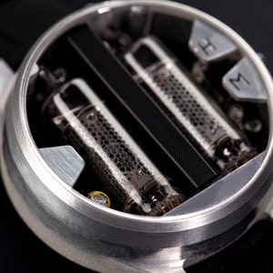 May include: Close-up of a unique wristwatch with a silver case. The watch face features two glass tubes with a honeycomb pattern and a black band. The watch has silver markers for hours and minutes.