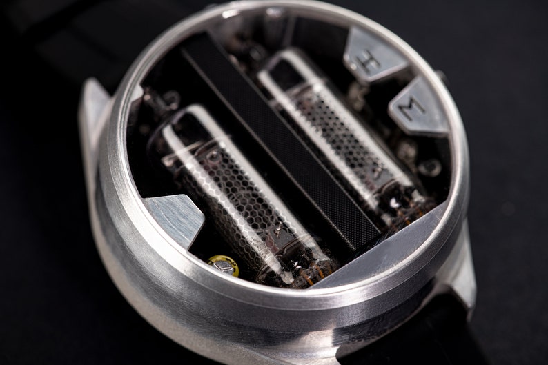 May include: Close-up of a unique wristwatch with a silver case. The watch face features two glass tubes with a honeycomb pattern and a black band. The watch has silver markers for hours and minutes.