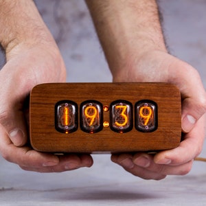 Puede incluir: Un reloj de madera con una pantalla de estilo vintage que muestra la hora 19:39. El reloj tiene cuatro tubos Nixie que se iluminan con números naranjas.