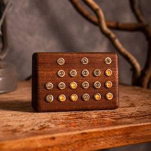May include: A rectangular wooden box with multiple brass-colored switches and small, illuminated orange lights. The box is made of dark wood and sits on a wooden surface. The background is a neutral gray.