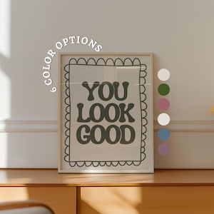 以下が含まれることがあります： レトロなフォントで「YOU LOOK GOOD」と書かれたフレーム入りのプリント。プリントは、スカラップのボーダーが付いた明るい色のフレームに入っています。6つのカラーオプションがフレームの左側に表示されています。