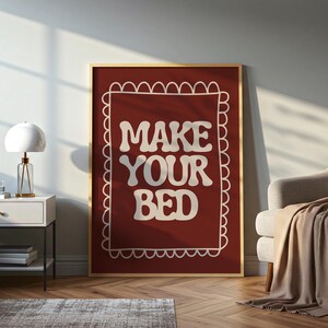 Könnte beinhalten: Ein gerahmter Wandkunstdruck mit den Worten "MAKE YOUR BED" in weißer, fetter Schrift im Retro-Stil auf einem tiefroten Hintergrund. Der Druck ist in einem goldfarbenen Rahmen gefasst und hat einen dekorativen weißen Rand.