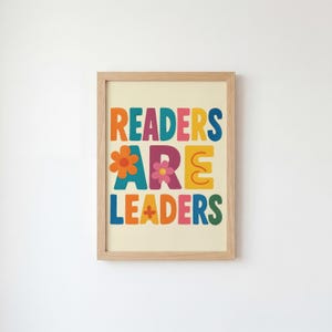 Könnte beinhalten: Ein gerahmter Druck mit den Worten "READERS ARE LEADERS" in farbenfrohen Buchstaben im Retro-Stil. Die Buchstaben sind orange, blau, gelb, lila und grün, mit Blumenakzenten. Der Rahmen hat eine helle Holzfarbe, und der Hintergrund ist cremefarben.