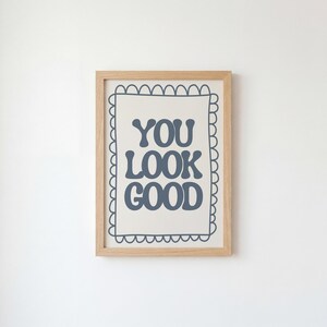Può includere: Una stampa incorniciata con la scritta "YOU LOOK GOOD" in blu scuro. Il testo è centrato su uno sfondo color crema, racchiuso da un bordo smerlato e una cornice in legno marrone chiaro. L'opera è esposta su una parete bianca.