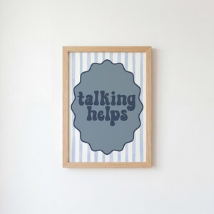 May include: A framed print with the words "talking helps" in a blue, wavy font. The text is inside a gray, scalloped shape, set against a background of blue and white vertical stripes. The frame is a light brown color.