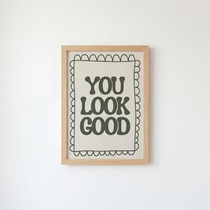 May include: A framed print with the words "YOU LOOK GOOD" in dark green, bold, retro-style lettering. The print is set against a light beige background and framed in a light brown wooden frame. The frame is hanging on a white wall.