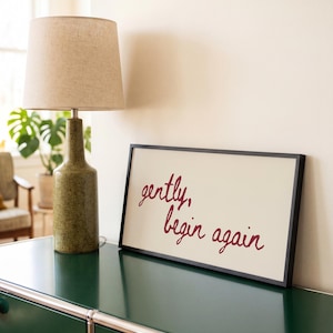 May include: A rectangular framed sign with a cream-colored background and the dark red cursive text "gently, begin again" displayed in the center. The sign is positioned next to a textured olive green lamp with a beige fabric lampshade.