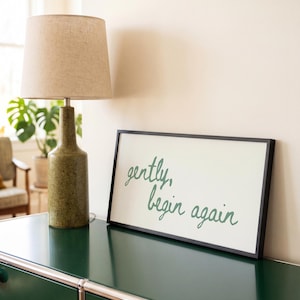 May include: A rectangular wall sign with a thin black frame and a white background featuring the green cursive text "gently, begin again" displayed on a dark green surface next to a textured green lamp with a beige fabric lampshade.