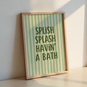 May include: A framed print with a light wood frame. The artwork features a striped background in light green and white, with the words "SPLISH SPLASH HAVIN' A BATH" in dark green lettering. The print is leaning against a white wall.