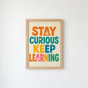May include: A framed print with the words "STAY CURIOUS KEEP LEARNING" in orange, teal, yellow, blue, pink, and green. The text is on a cream background, and the frame is light brown. The print has a cheerful, educational theme.