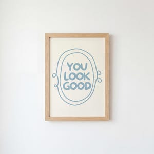 Può includere: Una stampa incorniciata con le parole "YOU LOOK GOOD" in blu all'interno di un design ovale blu. La cornice è in legno chiaro e lo sfondo è bianco sporco. La stampa è appesa a una parete bianca.