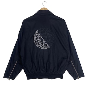 Puede incluir: Una chaqueta negra con un gran logotipo circular bordado de una cabeza de Medusa en la espalda. El diseño incluye un borde de greca, cuello clásico, mangas largas con puños de cremallera y un dobladillo acanalado sobre una percha de madera.
