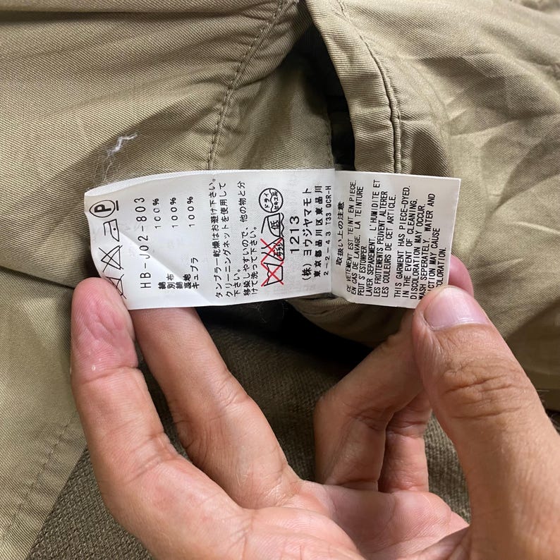 May include: Close-up of a clothing tag with Japanese text and care symbols. The tag is white with black text and includes fabric composition details. The garment appears to be a khaki-colored jacket or coat.