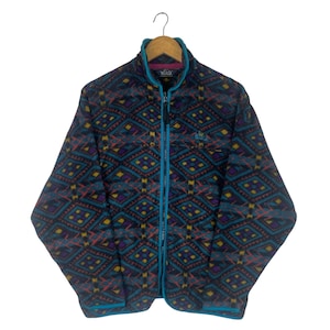 Op de afbeelding: Een vintage fleecejack met een geometrisch patroon in teal, paars, geel en rood. De jas heeft een turquoise ritssluiting over de hele lengte, turquoise biezen en een hoge kraag. De jas hangt aan een houten hanger.