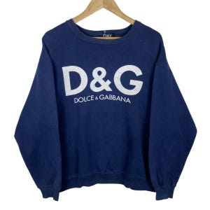 May include: A navy blue sweatshirt featuring the white text "D&G DOLCE & GABBANA". The long-sleeved crew neck top is suspended from a wooden hanger. The D&G logo is prominently displayed in white across the chest of the garment.