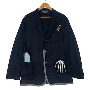 Pode incluir: Blazer preto com um patch de mão de esqueleto no bolso. O blazer tem uma borda cinza e um alfinete prateado com a letra "A" no bolso do peito. Um pequeno objeto azul está no outro bolso.