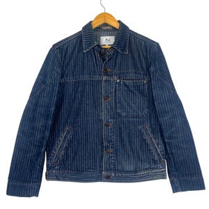 May include: A blue denim jacket with vertical white pinstripes. The jacket has a button-up front, two chest pockets, and two side pockets. The collar is a classic style, and the jacket is hanging on a wooden hanger. The label on the jacket reads "AJ".