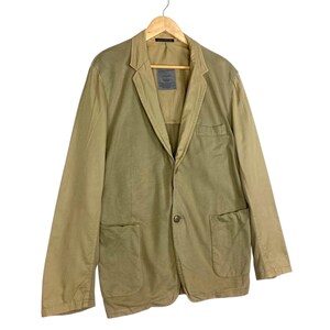 May include: A tan blazer with a two-button closure, two front pockets, and a notched lapel. The jacket is hanging on a wooden hanger. The blazer has a label on the inside of the collar.