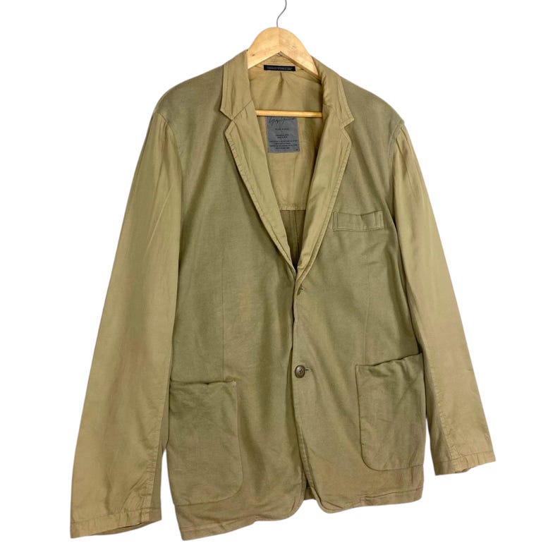 May include: A tan blazer with a two-button closure, two front pockets, and a notched lapel. The jacket is hanging on a wooden hanger. The blazer has a label on the inside of the collar.