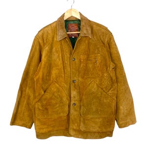 May include: A vintage, mustard-colored suede jacket with a corduroy collar. The jacket features two large front pockets, a chest pocket, and button closures. It is hanging on a wooden hanger.