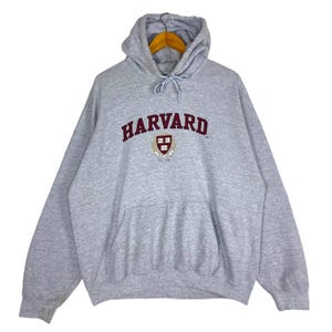 以下が含まれることがあります： ハーバード大学のグレーのフード付きスウェットシャツ。フロントポケットとドローストリングフード付き。「HARVARD」の文字が胸にアーチ状にマルーン色の文字で書かれており、下に大学の紋章があります。