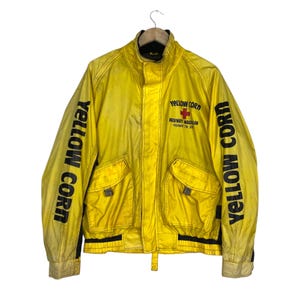 May include: A bright yellow jacket with black text reading YELLOW CORN printed vertically down both sleeves. The front features a red cross emblem above the text HIGHWAY MAGICIAN and YELLOW CORN. It has a high collar, two front pockets, and black trim.