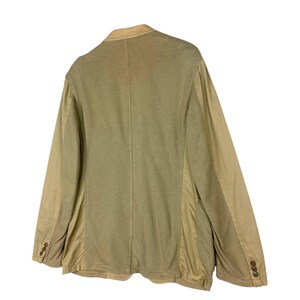 May include: A tan blazer with a two-button closure and a notched lapel. The jacket has a contrasting fabric on the sleeves and sides. The back of the blazer is visible, showing a center seam. The blazer is made of a soft, textured fabric.