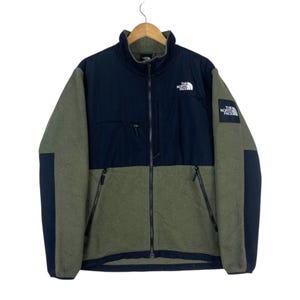 Può includere: Una giacca in pile con zip integrale caratterizzata da un design color-block in verde oliva e nero. La giacca presenta un colletto alto, due tasche laterali con zip, una tasca sul petto con zip e il logo The North Face sul petto.