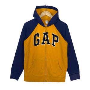 Könnte beinhalten: Ein senfgelber und marineblauer Kapuzenpullover mit Reißverschluss, langen Raglanärmeln und Kapuze. Das Wort GAP ist in marineblauen Buchstaben mit weißem Rand auf der Brust appliziert. Das Kleidungsstück hat einen Reißverschluss und zwei Taschen.