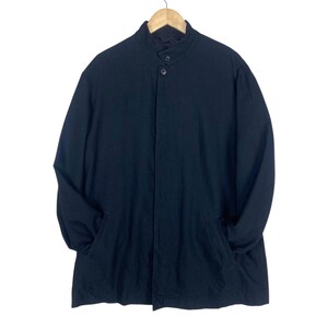 K&ouml;nnte beinhalten: Ein schwarzer langer Mantel mit Stehkragen und zwei Vordertaschen. Der Mantel h&auml;ngt an einem Holzb&uuml;gel vor wei&szlig;em Hintergrund. Der Mantel hat einen Knopfverschluss.