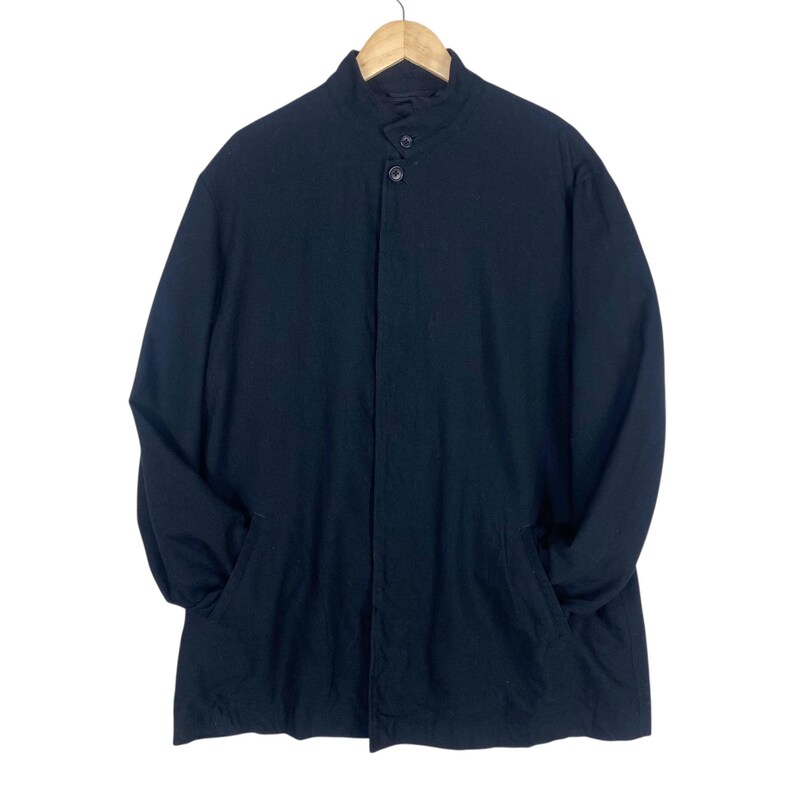 K&ouml;nnte beinhalten: Ein schwarzer langer Mantel mit Stehkragen und zwei Vordertaschen. Der Mantel h&auml;ngt an einem Holzb&uuml;gel vor wei&szlig;em Hintergrund. Der Mantel hat einen Knopfverschluss.