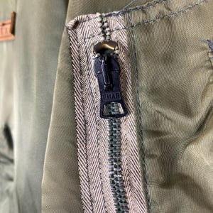 Puede incluir: Primer plano de una chaqueta verde oliva con cremallera negra. La cremallera est&aacute; cerrada y tiene la palabra "EMAR" impresa. Se aprecia el tejido de la chaqueta.