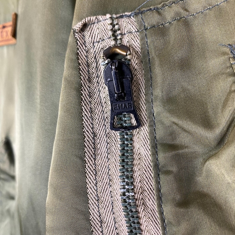 Puede incluir: Primer plano de una chaqueta verde oliva con cremallera negra. La cremallera est&aacute; cerrada y tiene la palabra "EMAR" impresa. Se aprecia el tejido de la chaqueta.