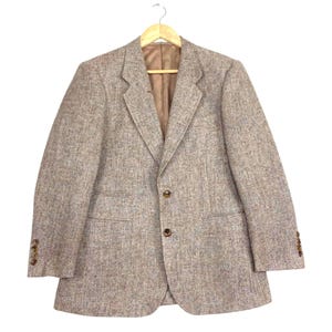 May include: A light brown tweed blazer featuring a textured speckled pattern, notched lapels, and a two-button front closure. The garment includes two front flap pockets, long sleeves with four buttons on each cuff, and a smooth brown interior lining.
