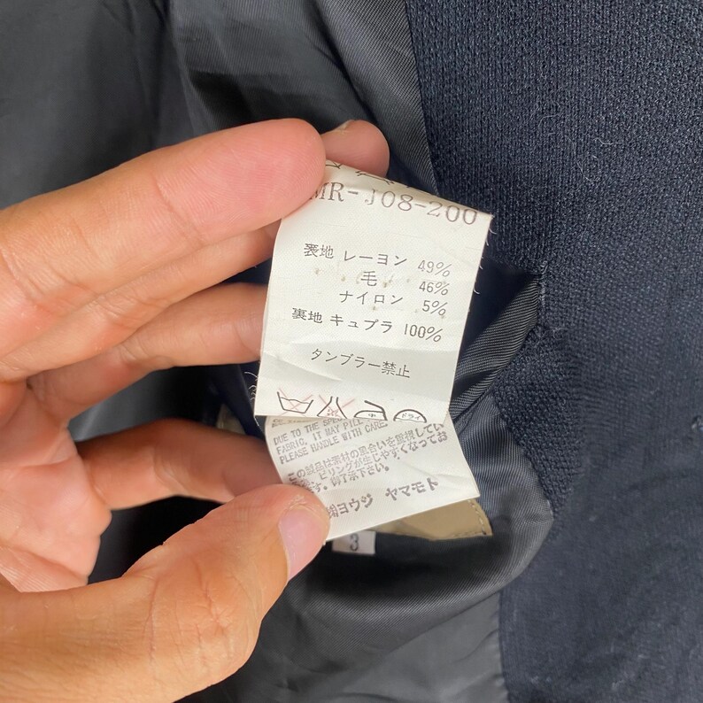 K&ouml;nnte beinhalten: Nahaufnahme eines Kleidungsst&uuml;cketiketts mit japanischem Text und Angaben zur Stoffzusammensetzung. Das Etikett listet die Prozents&auml;tze von Rayon, Wolle und Nylon sowie Pflegehinweise auf.