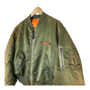 Puede incluir: Chaqueta bomber verde oliva con cierre de cremallera, dos bolsillos delanteros y un bolsillo peque&ntilde;o en la manga izquierda. La chaqueta tiene cuello, pu&ntilde;os y dobladillo de canal&eacute;. El forro interior es naranja. La marca "AVIREX LTD" es visible en el pecho.