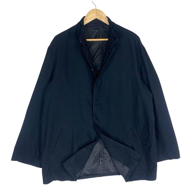 K&ouml;nnte beinhalten: Dunkelmarineblaue Jacke mit Knopfleiste und Kragen. Die Jacke h&auml;ngt an einem Holzb&uuml;gel. Die Jacke hat zwei Vordertaschen und ein schwarzes Futter. Die Jacke scheint aus einem schweren Stoff zu sein.