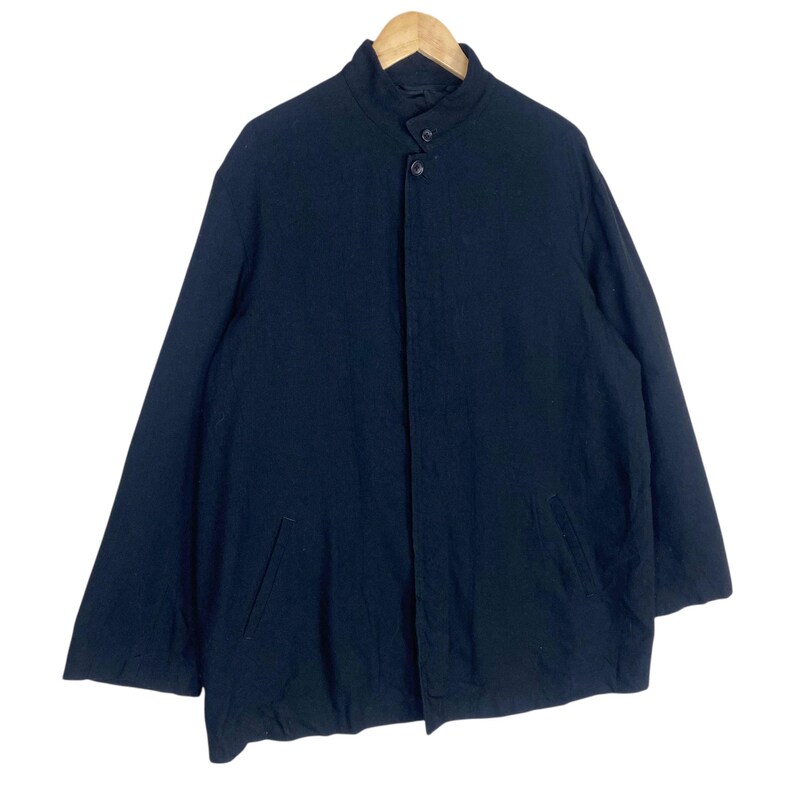 K&ouml;nnte beinhalten: Eine schwarze Jacke mit hohem Kragen und zwei Vordertaschen. Die Jacke hat einen Knopfverschluss und lange &Auml;rmel. Das Kleidungsst&uuml;ck h&auml;ngt an einem Holzb&uuml;gel vor wei&szlig;em Hintergrund.