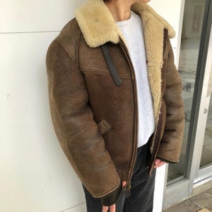 May include: A brown leather jacket with a cream-colored shearling collar and lining. The jacket has a zipper closure, a small pocket, and a strap detail on the collar. The sleeves have a dark green ribbed cuff.