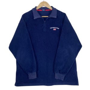 May include: Navy blue fleece pullover with a quarter-zip closure and a contrasting navy blue collar and cuffs. The shirt features the "POLO RALPH LAUREN" logo and a small American flag emblem on the chest. The long-sleeved shirt is hanging on a wooden hanger.