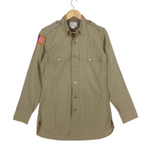 May include: A long-sleeve tan button-down shirt featuring two front chest pockets with button flaps, shoulder epaulets, and a small rectangular patch of the United States flag on the right sleeve. The shirt has a classic collar and buttoned cuffs.