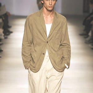 May include: A tan blazer with a single button closure, paired with cream-colored pants and a white undershirt. The outfit is worn by a person on a runway. The blazer has a classic, tailored fit.